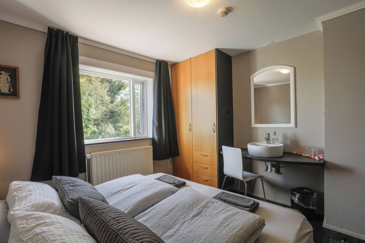 A bedroom with a large bed, a wooden wardrobe, a bathroom with a sink and mirror, and a window with curtains.