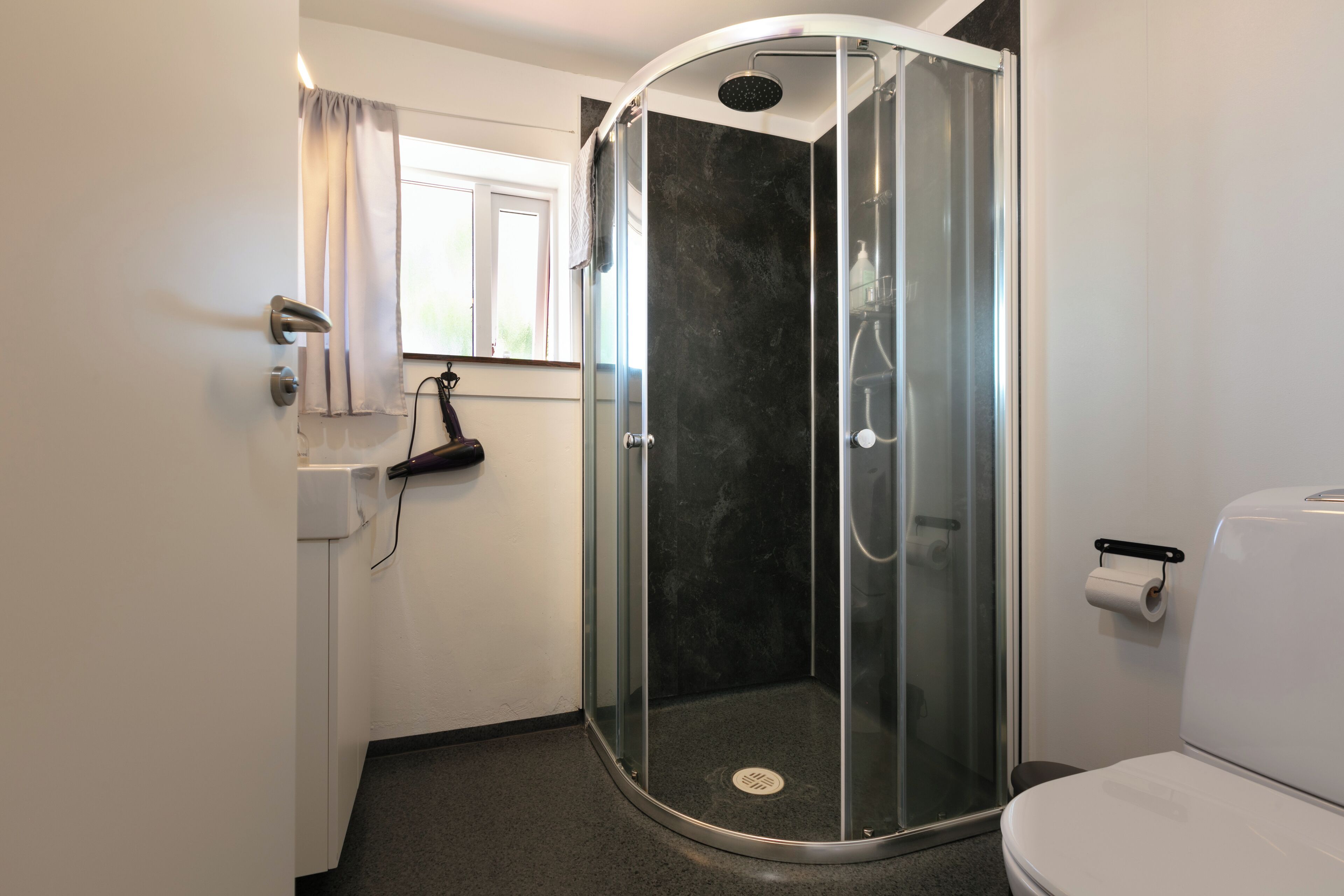 A modern bathroom with a glass-enclosed shower, a toilet, and a window with curtains.