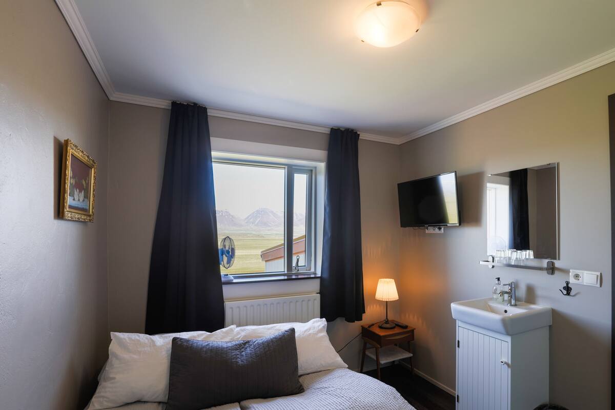 A bedroom with a bed, a window with curtains, a bedside lamp, a TV, a mirror, and a small cabinet.