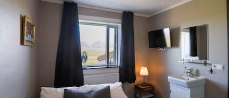 A bedroom with a bed, a window with curtains, a bedside lamp, a TV, a mirror, and a small cabinet.