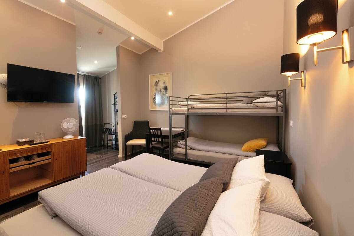 A hotel room with a large bed, a smaller bunk bed, a desk with a chair, a TV, and a wall-mounted lamp.