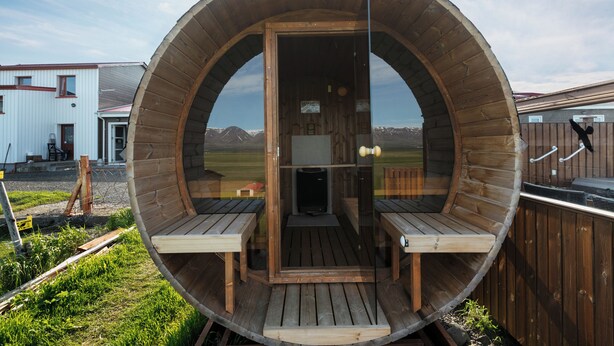 A wooden barrel-shaped structure with a door, a small table, and a bench inside. - Karuna Guesthouse (Skagafjörour)