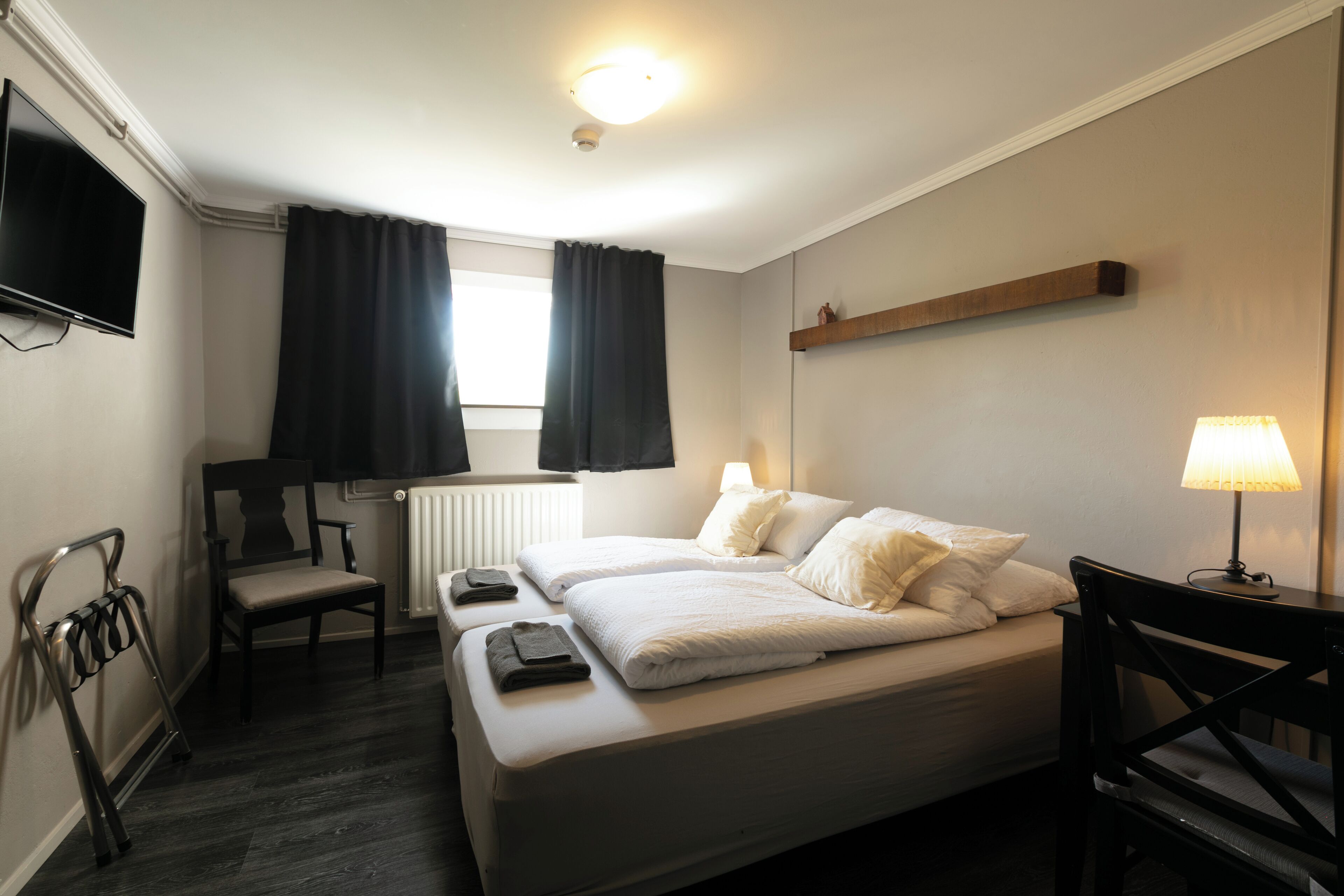 A hotel room with a bed, two chairs, a desk, a lamp, a TV, and a window with curtains.