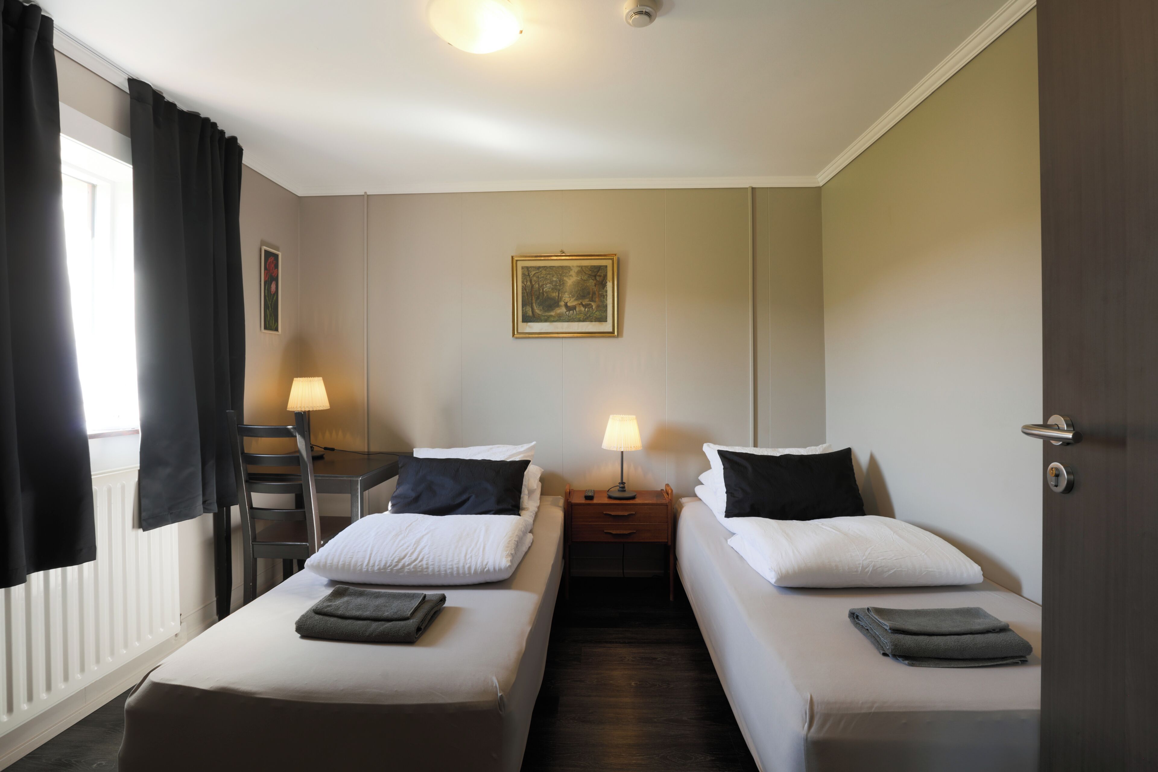 Hotellihuone, jossa on kaksi sänkyä, työpöytä, tuoli, lamppu ja taulu seinällä.