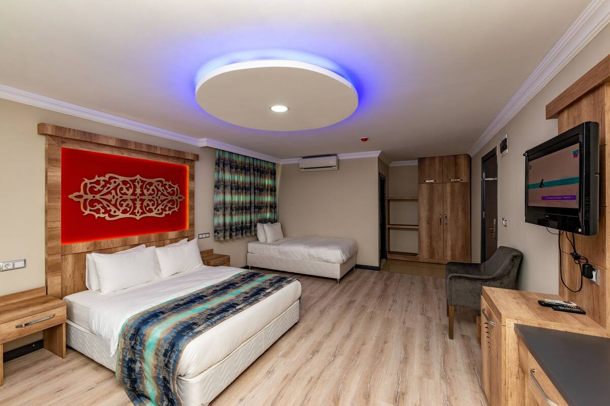 Quarto de hotel moderno com uma cama grande, uma cama menor, uma escrivaninha, uma televisão e uma luminária no teto.
