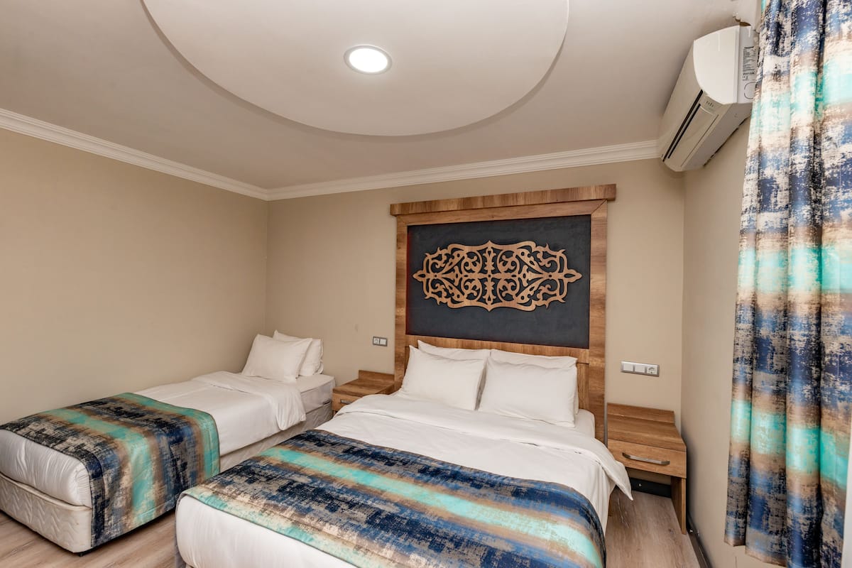Quarto de hotel com duas camas, uma cabeceira decorativa, uma pequena mesa de cabeceira de madeira e um ar condicionado.