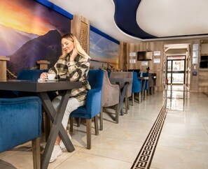 A person seated at a table in a hotel lobby with a mountainous mural on the wall, a modern ceiling design, and a patterned floor. - Linda Expo Palace (Antalya)