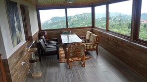 A dining area with a table and chairs, a painting on the wall, and a view of a hilly landscape through large windows.