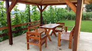 A wooden gazebo with a table and chairs, surrounded by lush greenery and a view of a garden.
