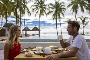 Um casal jantando em uma mesa de madeira com itens de café da manhã, com vista para uma piscina e palmeiras.