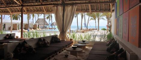 A beachside lounge with thatched roof, cushioned seating, and a view of the ocean.