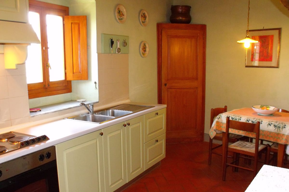 Een keuken met witte kastjes, een spoelbak en een fornuis. Er staat een eettafel met stoelen en een kom op.
