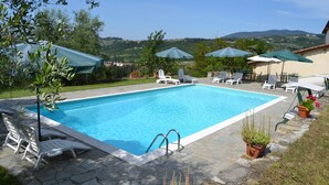A rectangular swimming pool with a stone deck, surrounded by white lounge chairs and umbrellas, with a view of hills in the background. - Agriturismo Montereggi (Fiesole)