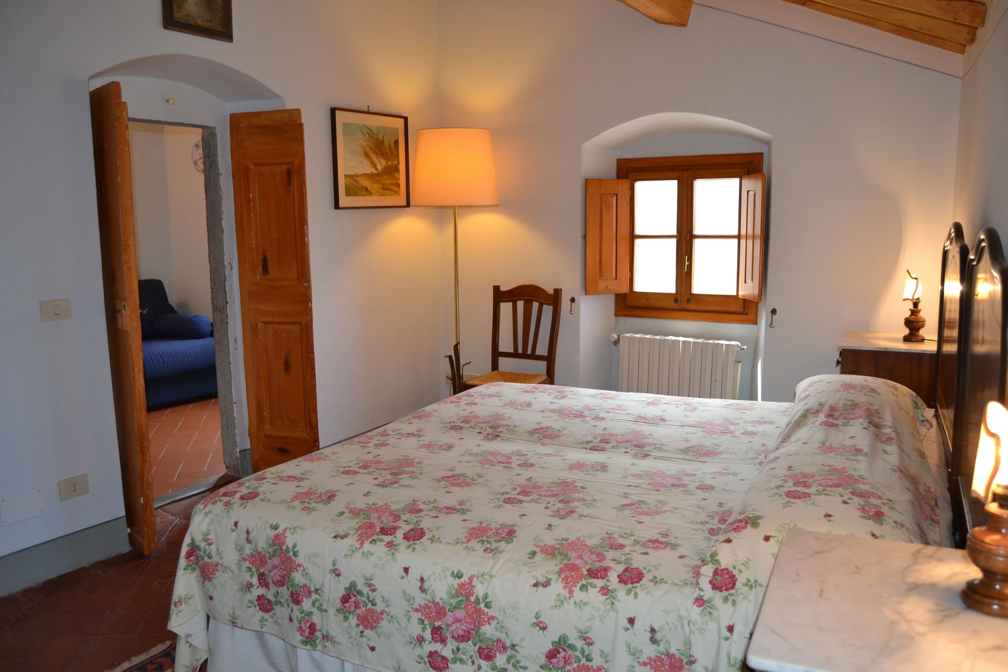 Un dormitorio con colcha estampada, una silla de madera, una mesita de noche con lámpara y una ventana con contraventanas de madera.