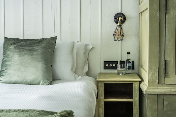 Una cama bien hecha con un almohadón verde y ropa de cama blanca, una mesita de noche con una botella y vasos, y una lámpara de pared.