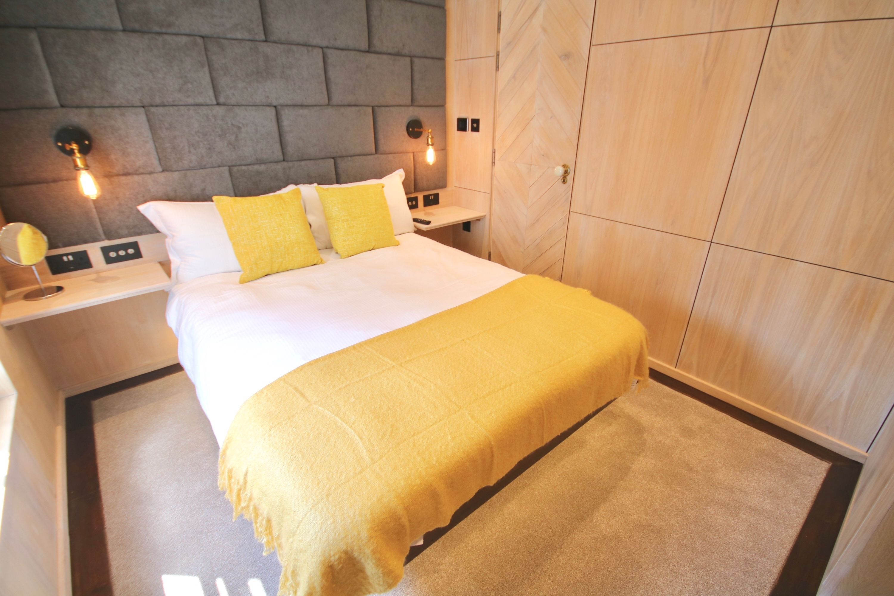 A neatly made bed with a yellow throw, a bedside table with a lamp, and a wardrobe with a herringbone pattern.