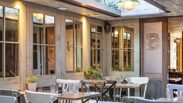 Restaurante com mesas ao ar livre, janelas amplas e decoração rústica.