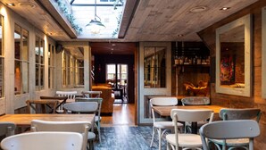 Interior de restaurante com mesas e cadeiras de madeira, área de bar com prateleiras e vista para a rua através de grandes janelas.