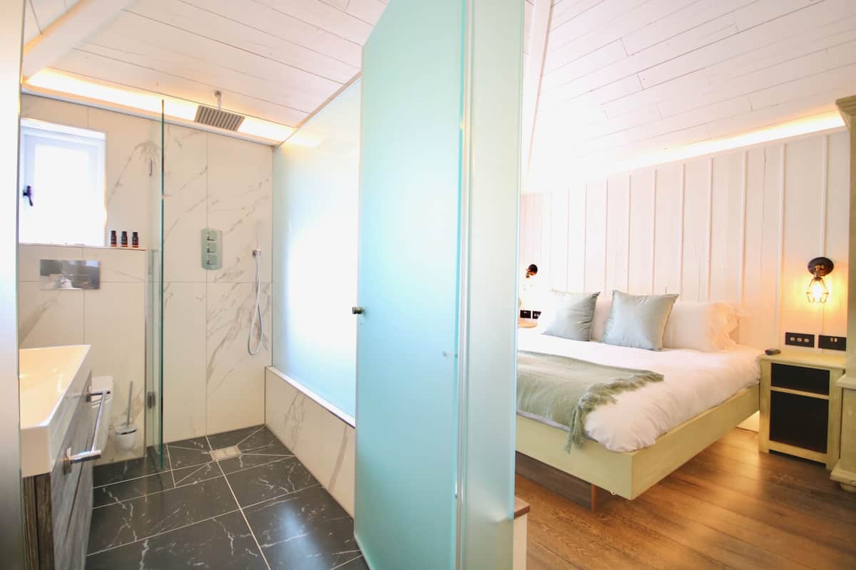 A modern bedroom with a bed, bedside tables, and a nightstand. There is a bathroom with a shower, sink, and toilet.