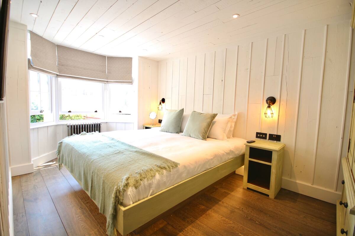 A bedroom with a large bed, wooden flooring, a nightstand with a lamp, and a window with a view of greenery.