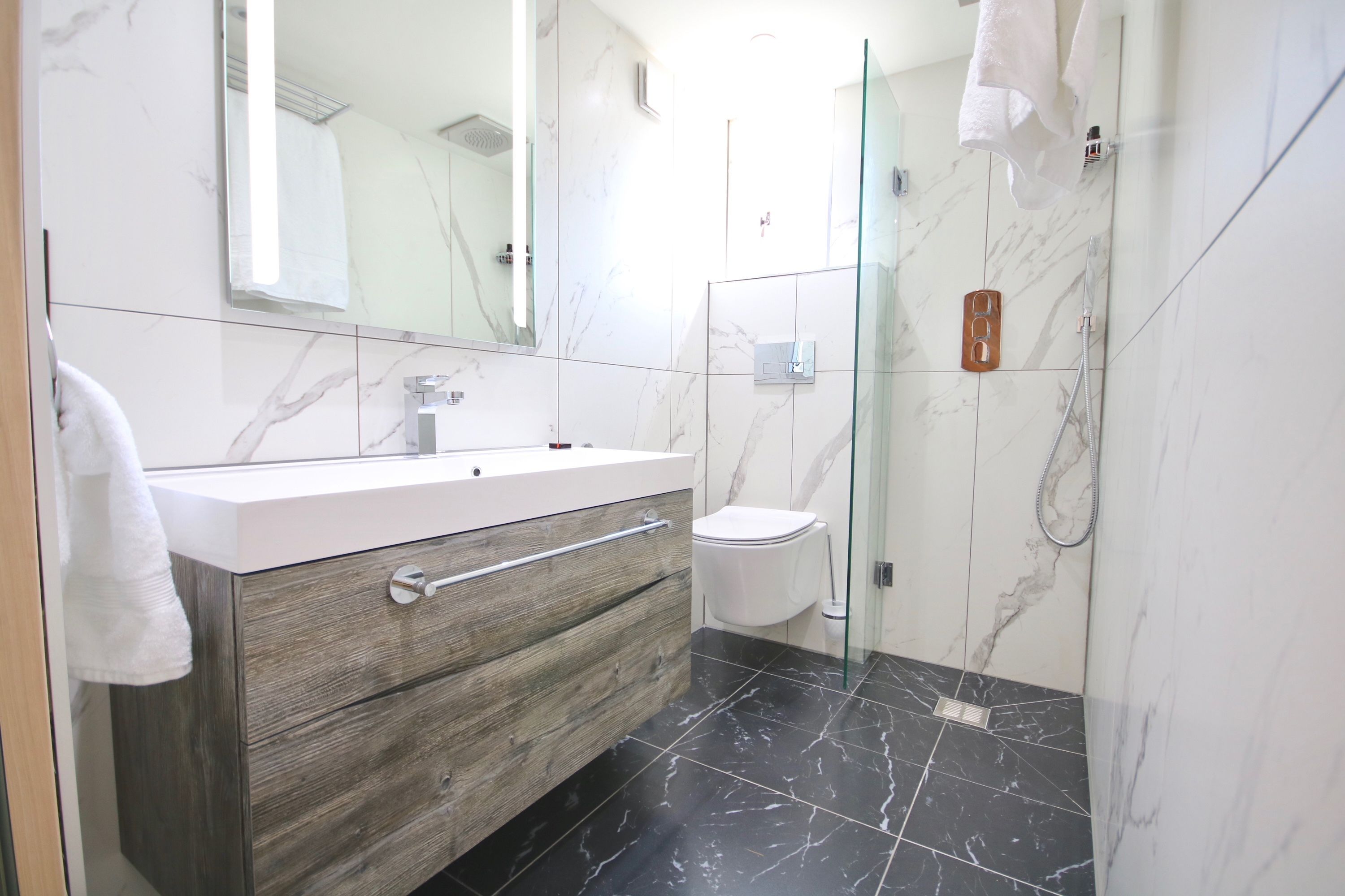 Un baño moderno con piso de mármol, tocador de madera, cabina de ducha de vidrio y un inodoro blanco.