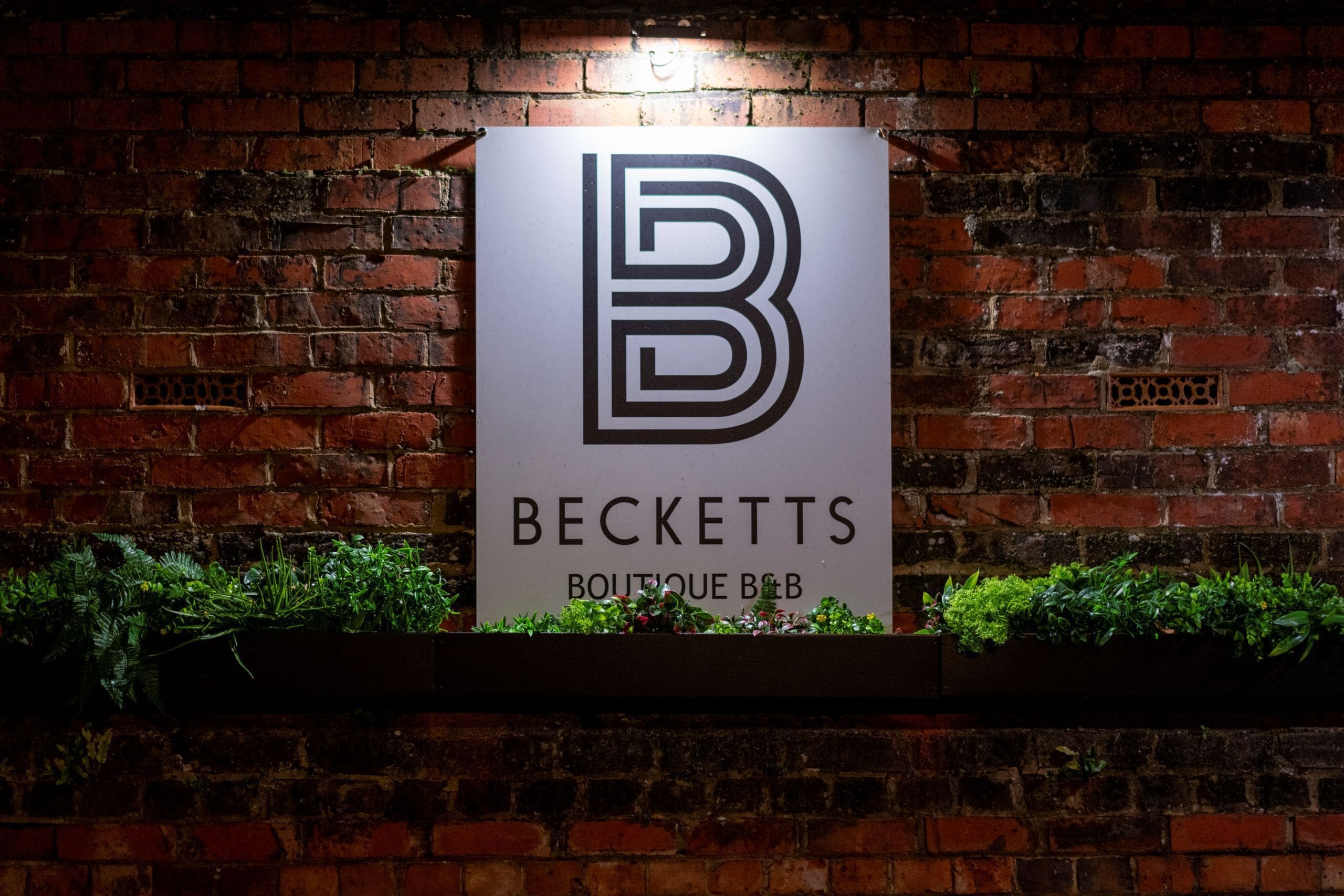 A sign for 'BECKETTS BOUTIQUE B&B' mounted on a brick wall with a planter below.