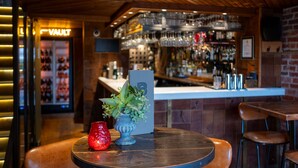 Uma área de bar com teto de madeira, um balcão com vários copos e uma placa com a inscrição 'VAULT'.