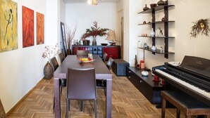 Zona pranzo con un lungo tavolo in legno, sedie e un pianoforte. Sono presenti opere d'arte alle pareti e uno scaffale con oggetti decorativi.