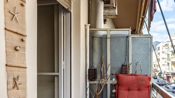 Un balcone con una sedia di legno, una poltrona imbottita, un tavolo con delle tazze, una pianta in vaso e una finestra con un condizionatore d'aria.