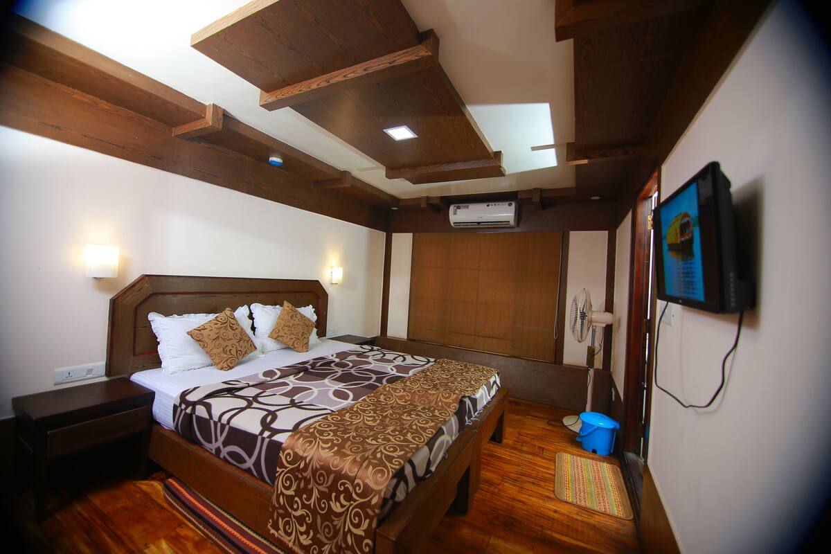 A hotel room with a wooden bed, a nightstand, a wall-mounted air conditioner, a ceiling fan, and a television.