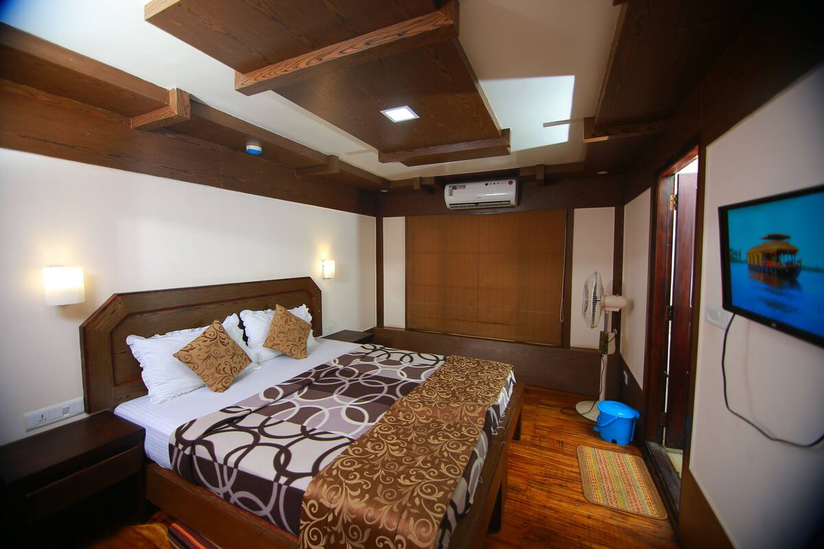 A bedroom with a wooden headboard, a bed with patterned bedding, a bedside table, a wall-mounted fan, a television, and a wooden floor.