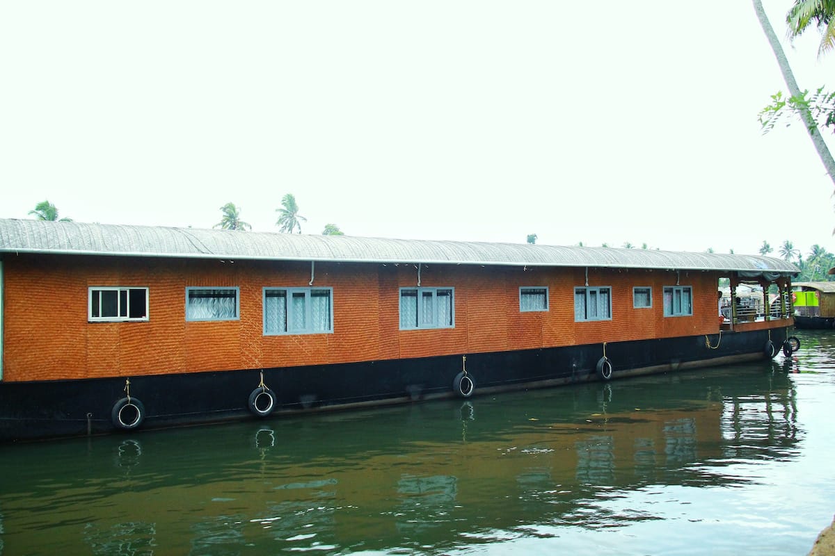 A long, orange and black houseboat with multiple windows and a flat roof, floating on calm water.