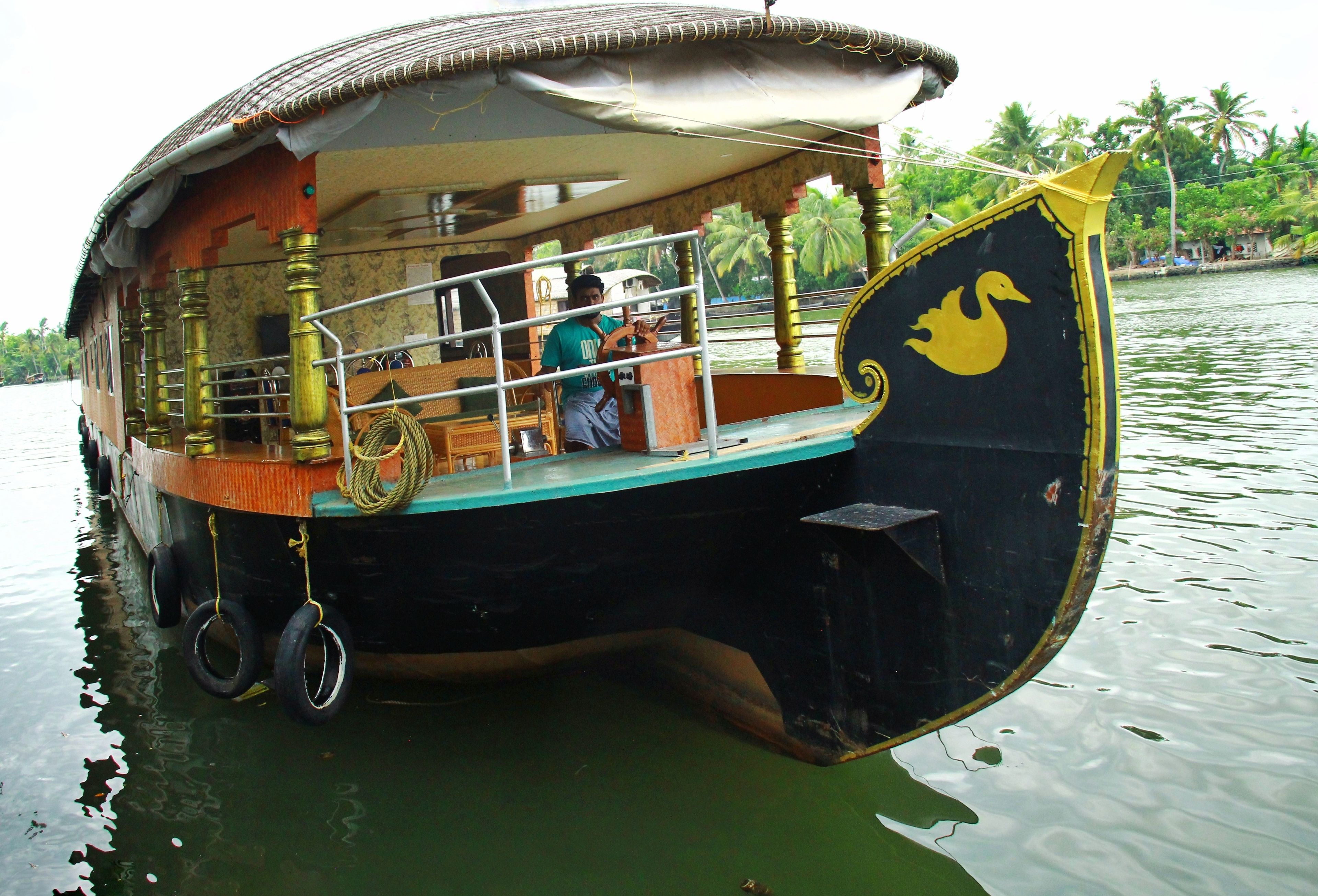 A houseboat with a thatched roof, a yellow swan design on the bow, and a person steering the boat.