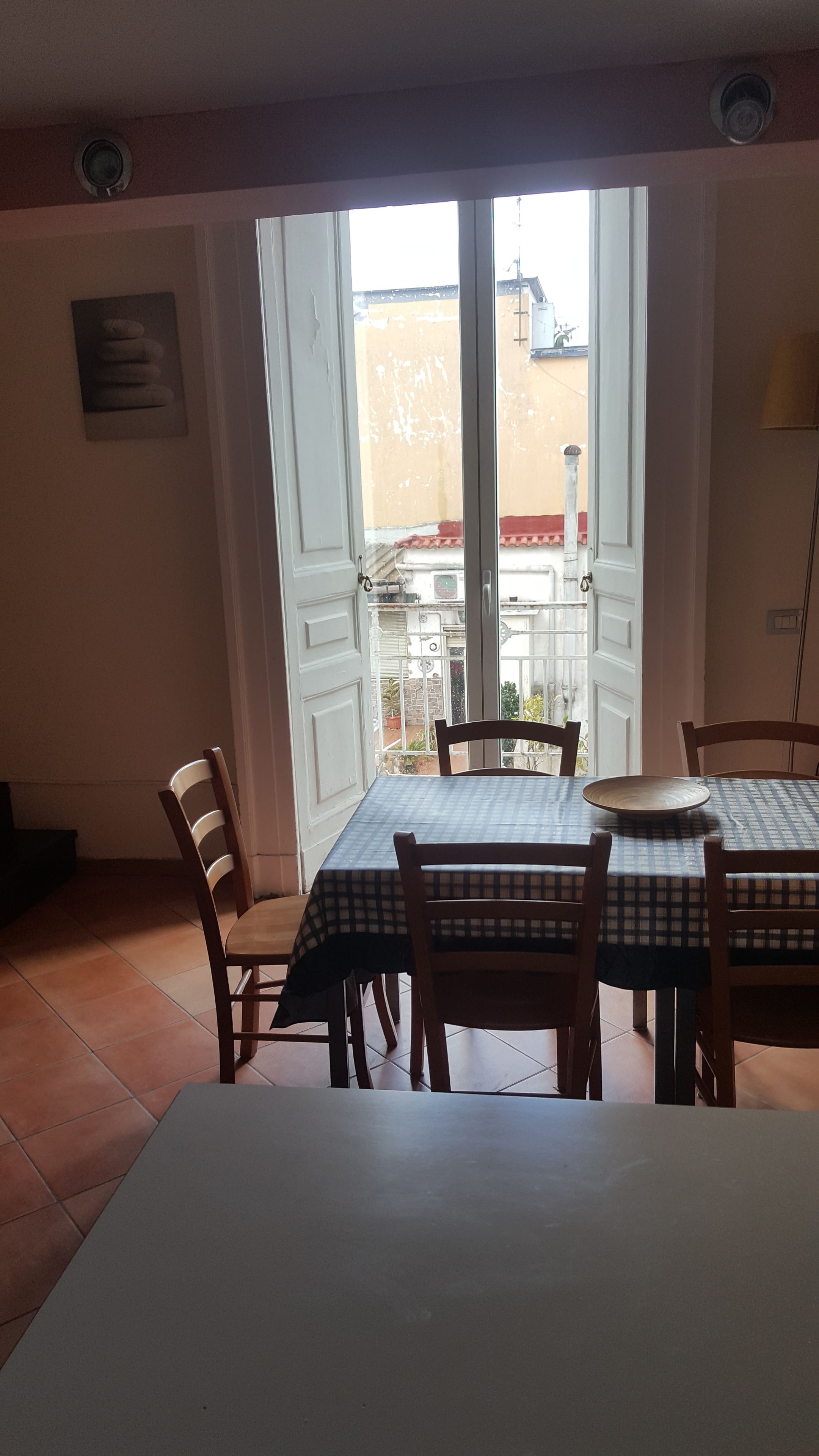 Zona pranzo con tavolo apparecchiato per quattro persone, sedie, vista su un balcone esterno tramite una porta in vetro e un lampadario a parete.