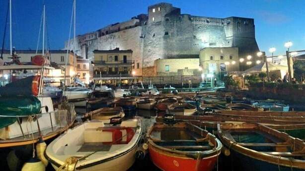 A harbor with boats docked, a historic building in the background, and a clear sky. - Miseria e Nobiltà (Naples)