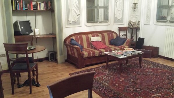 A living room with a sofa, a coffee table with magazines, a bookshelf, a computer, and a window with curtains. - Miseria e Nobiltà (Naples)