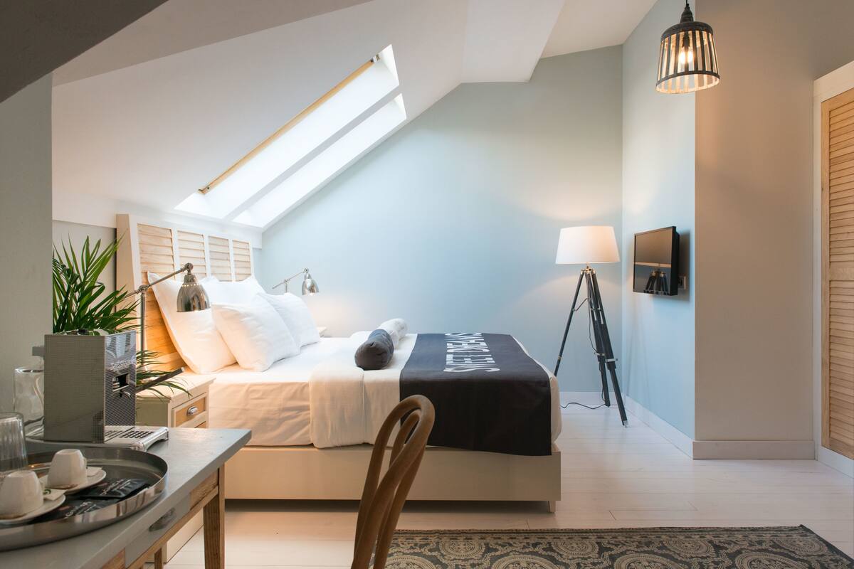 A modern bedroom with a skylight, a bed with a black cover, bedside lamps, a desk with a tray of cups, and a potted plant.