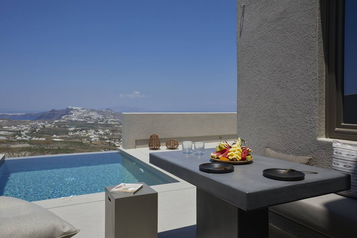 An infinity pool with a clear view of a distant town and mountain, a table with a fruit bowl, and a few glasses.