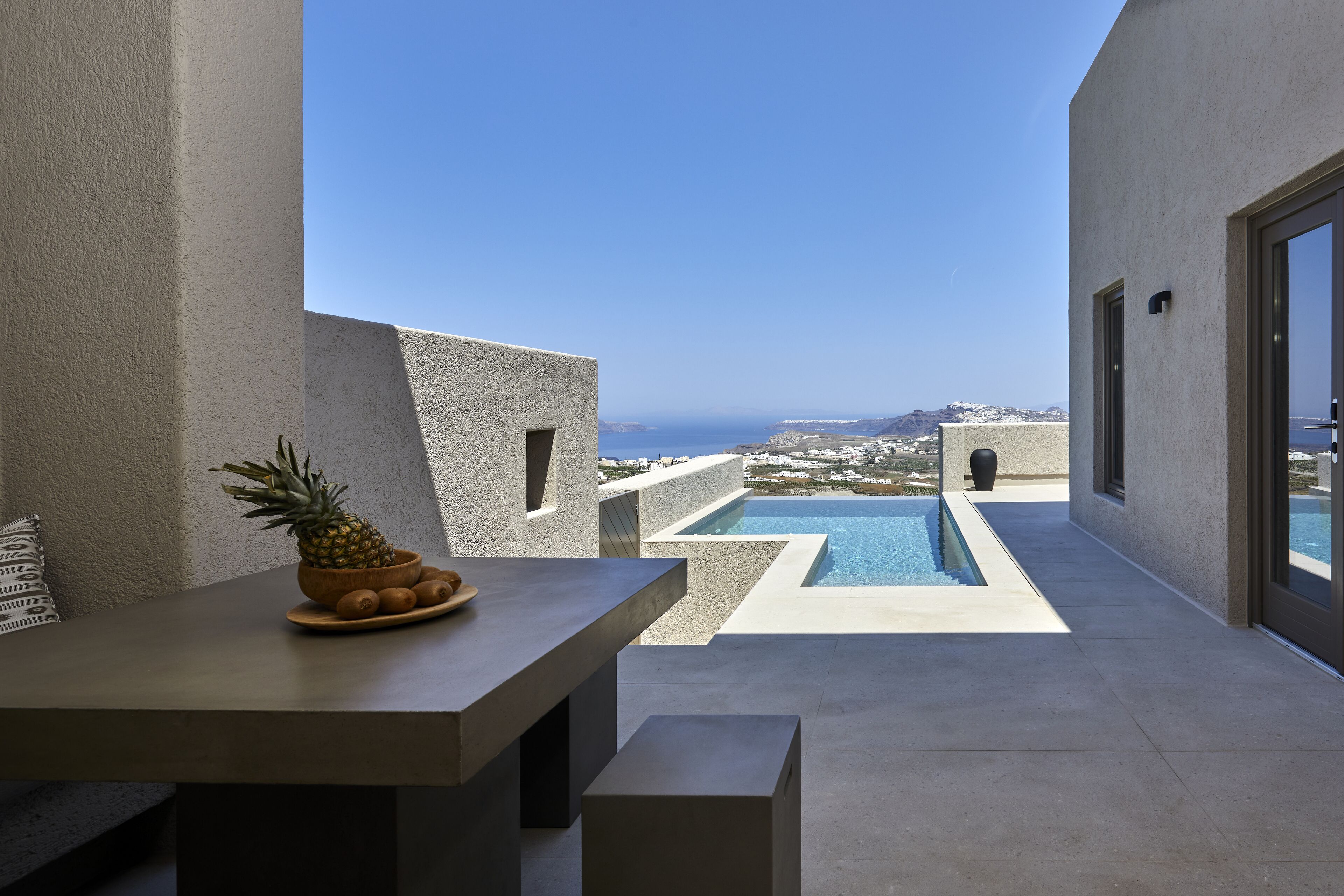 A modern outdoor terrace with a pool, a table with a pineapple centerpiece, and a view of the coastline.