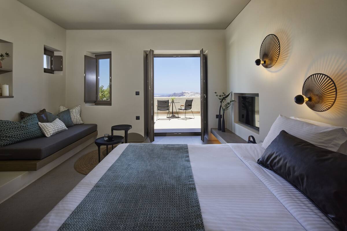A modern hotel room with a large bed, a sofa, a small table, and a balcony with a view.