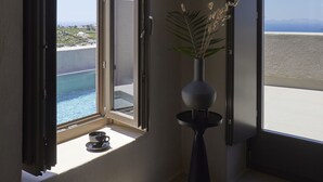 Una habitación con un ventanal grande que da a una piscina y vistas al mar. Hay una mesita con un libro y un jarrón con flores secas.