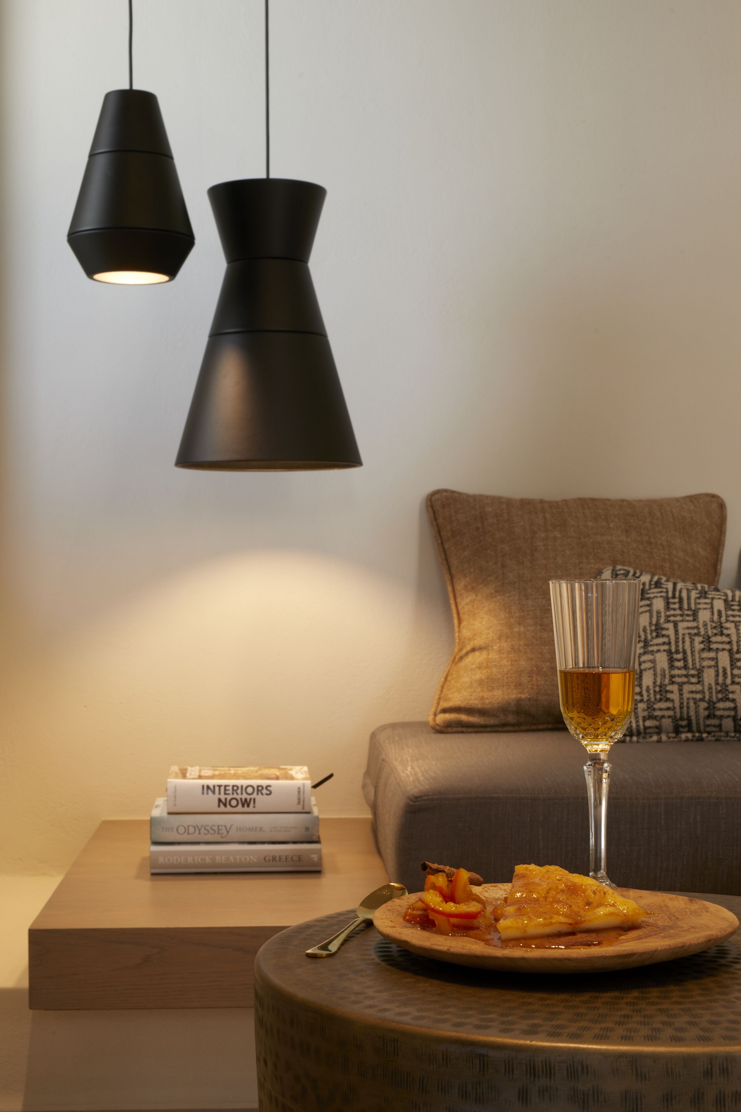 A modern living room with a sofa, a coffee table with a plate of food and a glass of wine, and two pendant lights. There are books on a small table beside the sofa.