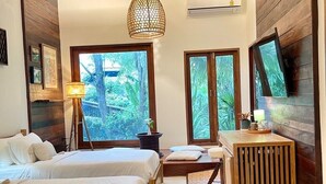 Un dormitorio con dos camas, un escritorio de madera, un tapete tejido y ventanas amplias con vista a follaje.