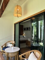 Quarto com cama, mesa, cadeiras, luminária de vime e vista para o verde através de uma porta de vidro.