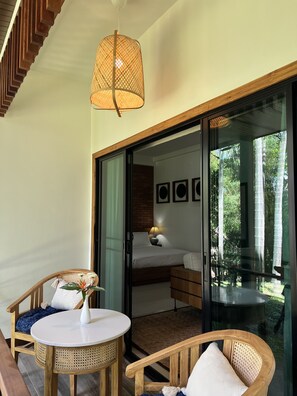 Una habitación con una cama, una mesa, sillas, una lámpara de mimbre y vistas a la vegetación a través de una puerta de cristal.