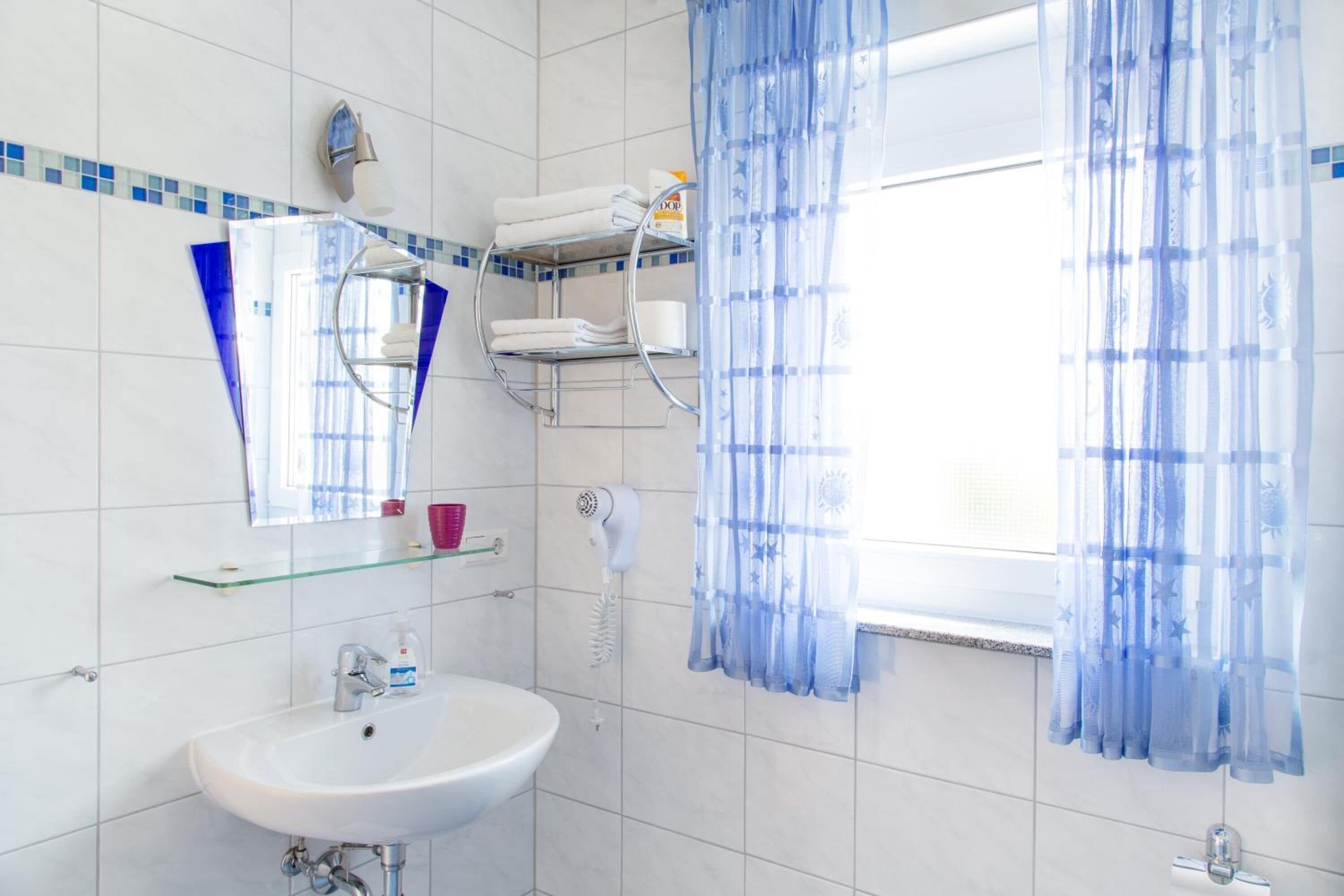 A bathroom with a white sink, a mirror, a towel rack, a hairdryer, and a window with blue curtains.