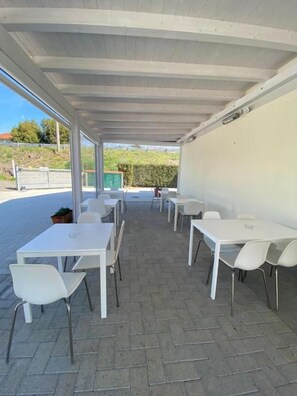 An outdoor covered patio with white tables and chairs, a paved floor, and a view of the surrounding area.
