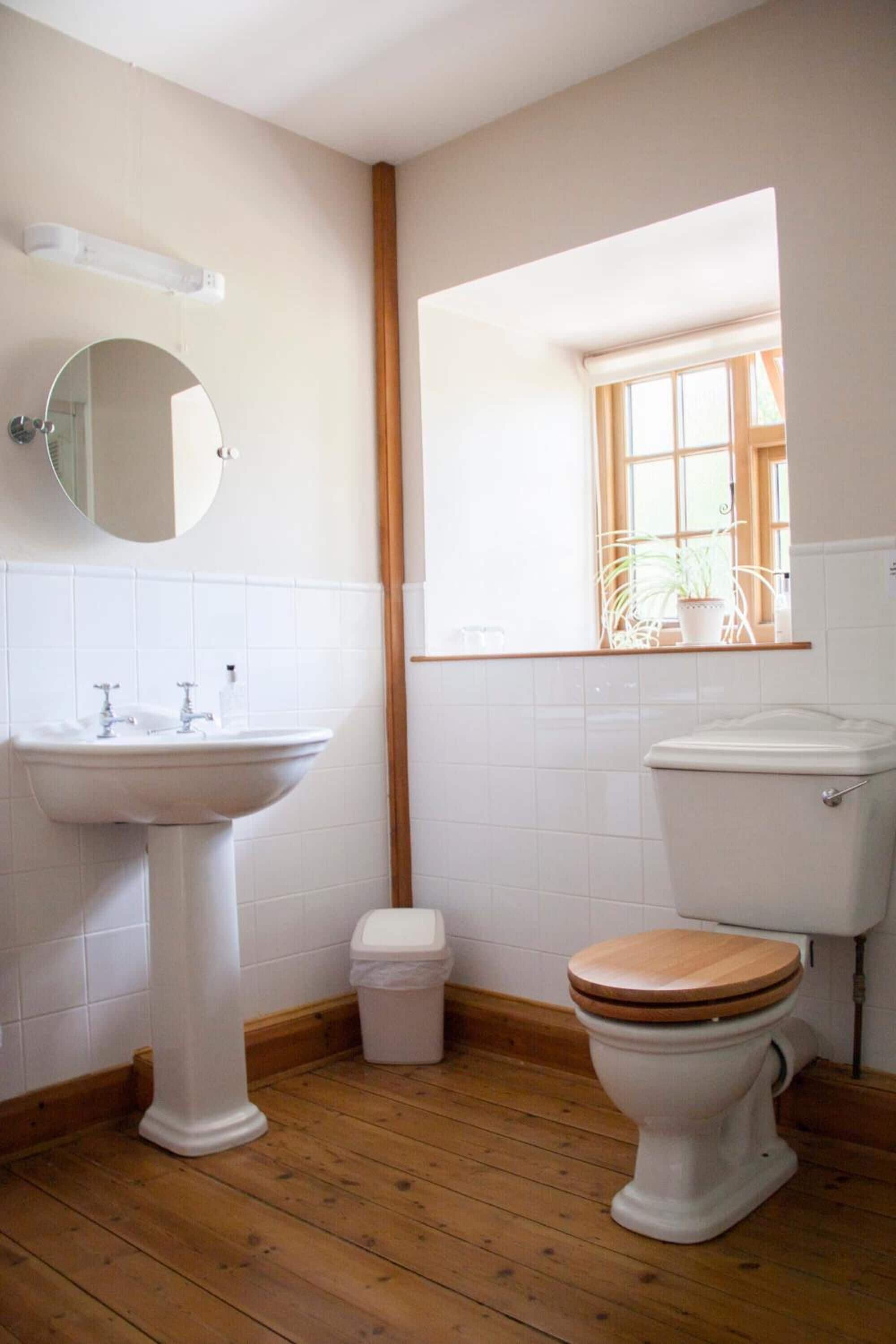Un baño con suelo de madera, azulejos blancos, un lavabo, un inodoro y una ventana con una planta.
