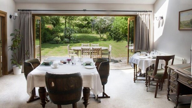 Comedor con una mesa preparada para cuatro personas, un piano y vistas a un jardín a través de un ventanal abierto.
