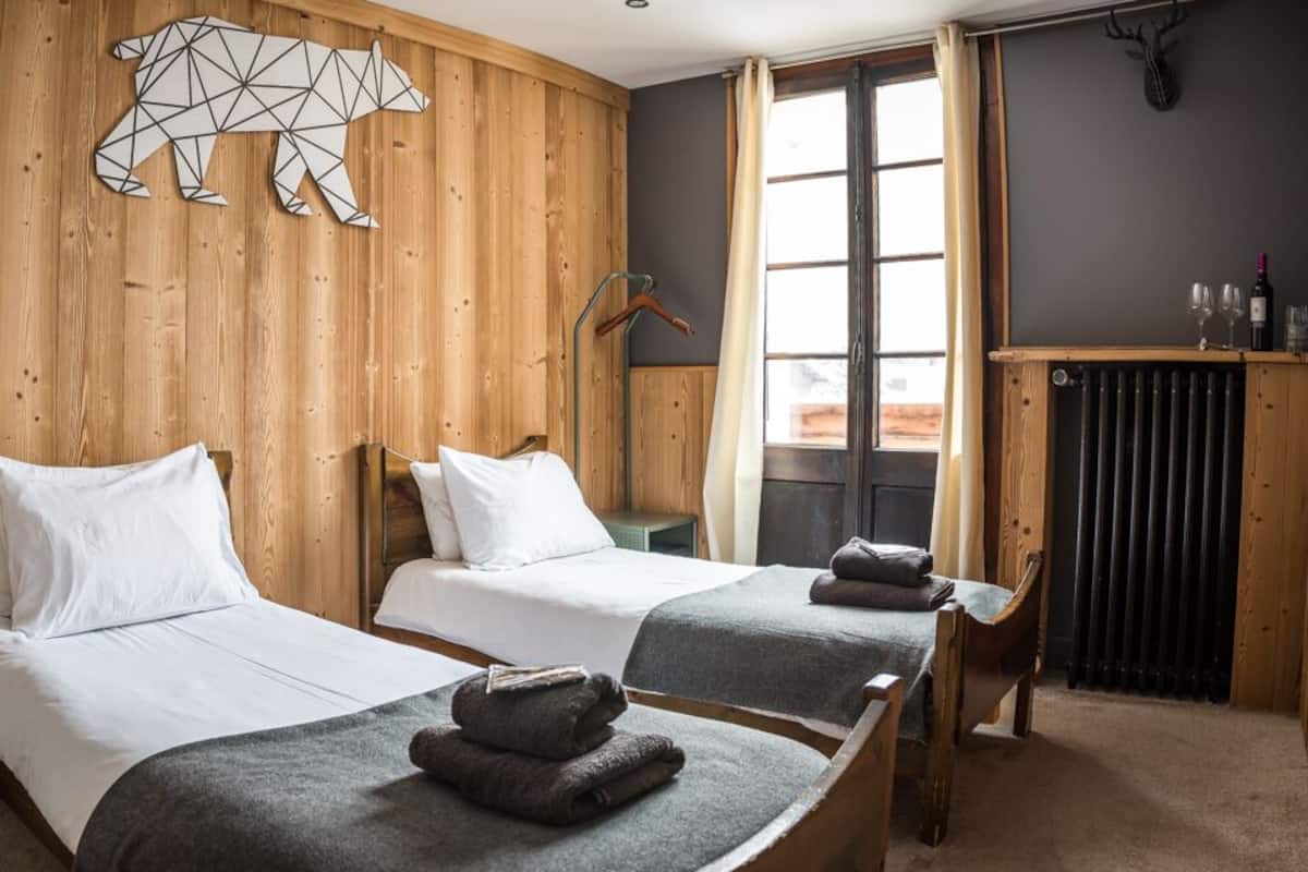 Una habitación con dos camas, paredes de madera, una ventana con cortinas, un radiador y una cabeza de venado colgada.
